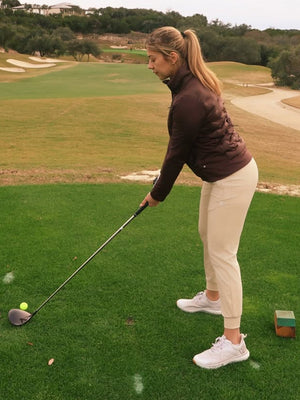 Model playing golf, wearing women's lightweight fleece-lined puffer jacket in coffee brown on the course.
